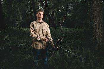 high school senior archery pose with bow and arrow in the natural woods environment using off camera lighting