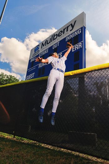 high school senior baseball player catching a fly ball over the fence in front of the score board.