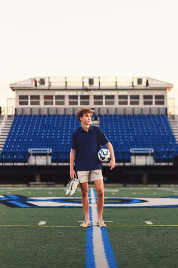 high school senior soccer player posing with shoes and ball on home soccer field at susnet